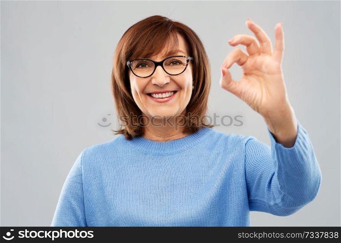 gesture, vision and old people concept - portrait of smiling senior woman in glasses showing ok hand sign over grey background. happy senior woman in glasses showing ok hand sign