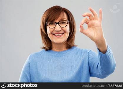 gesture, vision and old people concept - portrait of smiling senior woman in glasses showing ok hand sign over grey background. happy senior woman in glasses showing ok hand sign