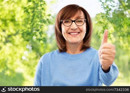 gesture, vision and old people concept - portrait of smiling senior woman in glasses showing thumbs up over green natural background. portrait of senior woman showing thumbs up