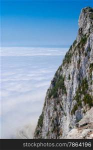 Gibraltar cliff face above clouds with road below against clear blue sky.