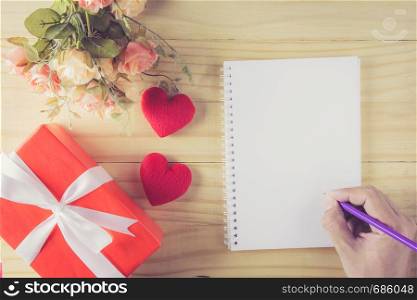 Gift box white bow ribbon and heart hand holding pencil with notebook on wood table background top view, february of valentine's day in concept.