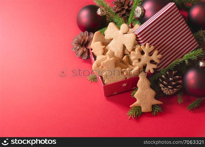 Gift box with honey and ginger Xmas cookies, red globes, pine cones, and fir twigs, on a red background. Christmas banner, Xmas baking, and gifting.