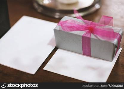 Gift box with pink ribbon and white blank greeting card paper mock-up on the reception table with copy space