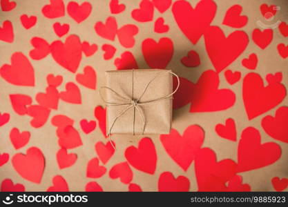 Gift box wrapped in kraft paper surrounded by red hearts on brown plain paper background, Valentines Day concept top view modern design, copy space romantic. Gift box wrapped in kraft paper surrounded by red hearts on brown plain paper background, Valentines Day concept top view modern design, copy space