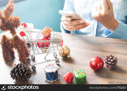 Gift boxes in shopping cart and Christmas decorations on table, Woman using mobile device, Online shopping concept
