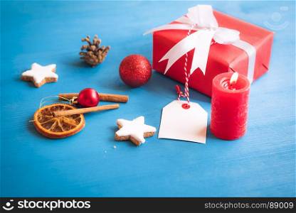 Gift wrapped in red paper and tied with white ribbon and bow, with an empty label attached to it, surrounded by star shaped cookies, spices, globes, and red candle.