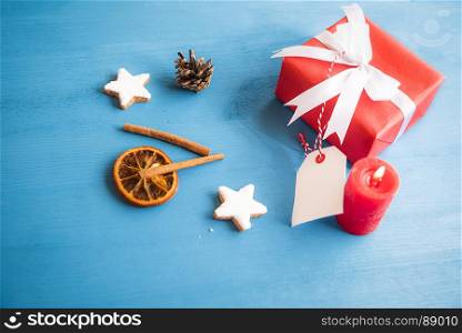 Gift, wrapped in red paper and tied with white ribbon and bow, with a blank tag attached to it, surrounded by cinnamon sticks, dried orange and star shaped cookies.