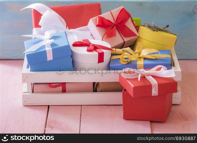 Gifting theme image with a bunch of colorfully wrapped presents, in a white wooden crater, on a pink table and a blue wooden background.