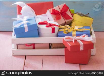 Gifting theme image with a bunch of colorfully wrapped presents, in a white wooden crater, on a pink table and a blue wooden background.
