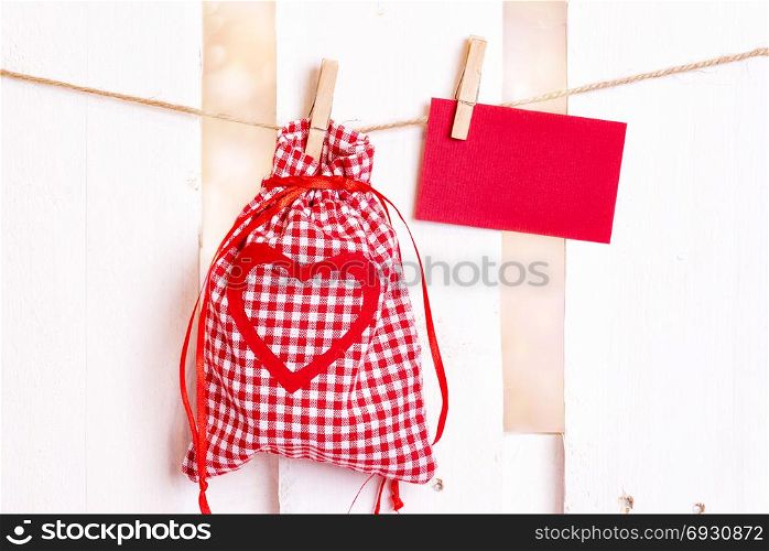 Gifting theme image with a checkered fabric pouch and a decorative red heart and an empty message card, tied to a string with wooden clips, on a white fence.