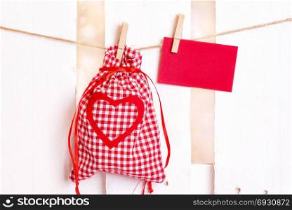 Gifting theme image with a checkered fabric pouch and a decorative red heart and an empty message card, tied to a string with wooden clips, on a white fence.