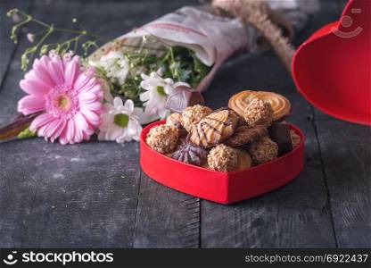 Gifting theme image with a red heart-shaped box full of treats, chocolate, cookies and a bouquet of flowers in the background, on a black wooden table.