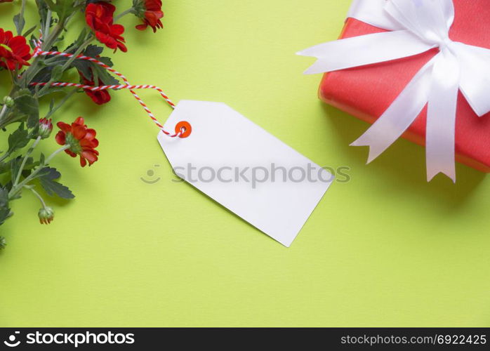Gifting theme image with lovely red flowers, an empty label tied to it and a red gift tied with white ribbon and bow, on a green background. Greeting card idea.