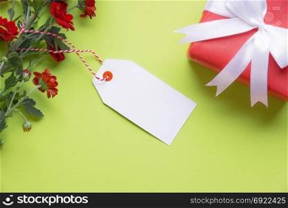 Gifting theme image with lovely red flowers, an empty label tied to it and a red gift tied with white ribbon and bow, on a green background. Greeting card idea.