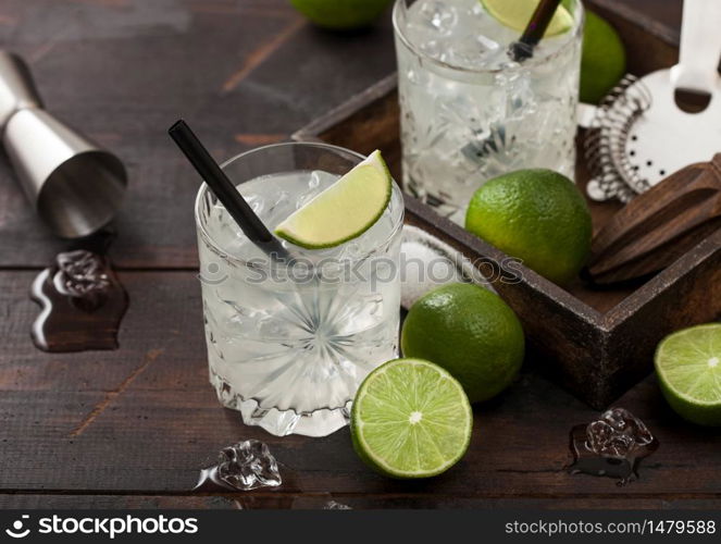 Gimlet Kamikaze cocktail in crystal glasses with lime slice and ice on wooden background with fresh limes and jigger with strainer.