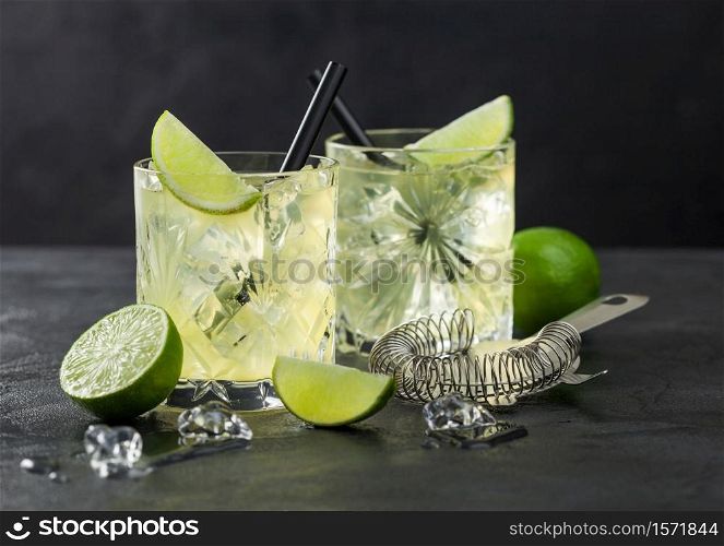 Gimlet Kamikaze cocktail in crystal glasses with lime slice and ice on black background with fresh limes and strainer.