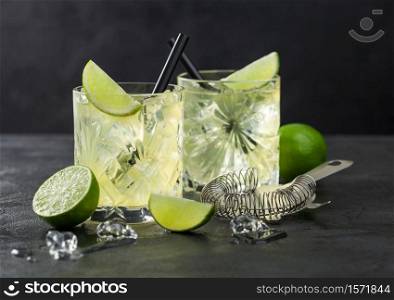 Gimlet Kamikaze cocktail in crystal glasses with lime slice and ice on black background with fresh limes and strainer.