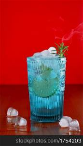 Gin and tonic, alcoholic drink with lime, rosemary and ice on rustic table, bright red background. Blue glass tumbler, smoke rises above the rosemary branch. Refreshing summer drink. Selective focus