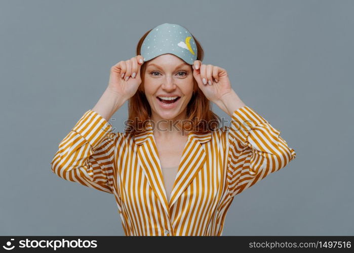 Ginger female model wears sleepmask and striped pajamas, awakes in good mood, realizes today is weekend, can stay in bed for long time, smiles positively, isolated on grey background. Sleeping