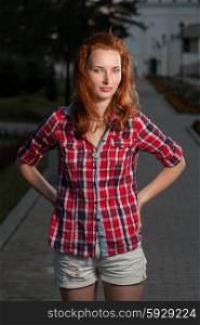 Ginger haired women in jeans shorts and red shirt posing outdoors. Torso shot front view