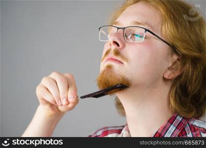 Ginger nerd man brushing his beard using comb taking care of facial hair.. Man brushing his beard using comb