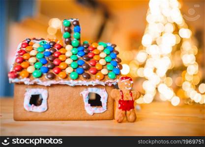 Gingerbread fairy house on a background of bright Christmas tree with garland. Homemade Christmas Gingerbread House on a table. Christmas tree lights in the background