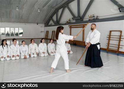 Girl and aikido master fighting with wooden sword bokken during martial arts class. Teenage female group sitting nearby on floor and watching. Girl and aikido master fighting with wooden sword during martial arts class