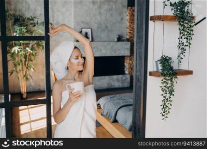 Girl applies anti-aging eye patches, smiling. Weekend morning at home. Lady with coffee relaxes in luxurious spa. Beautiful European in towel after bathing.