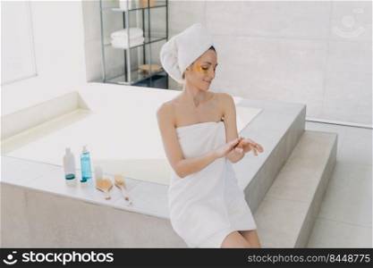 Girl applies eye patches and hand cream. Attractive caucasian woman wrapped in towel after bathing. Young hispanic lady takes shower at home. Relaxation at spa resort. Modern interior of bathroom.. Girl applies eye patches and hand cream. Attractive caucasian woman wrapped in towel at spa resort.