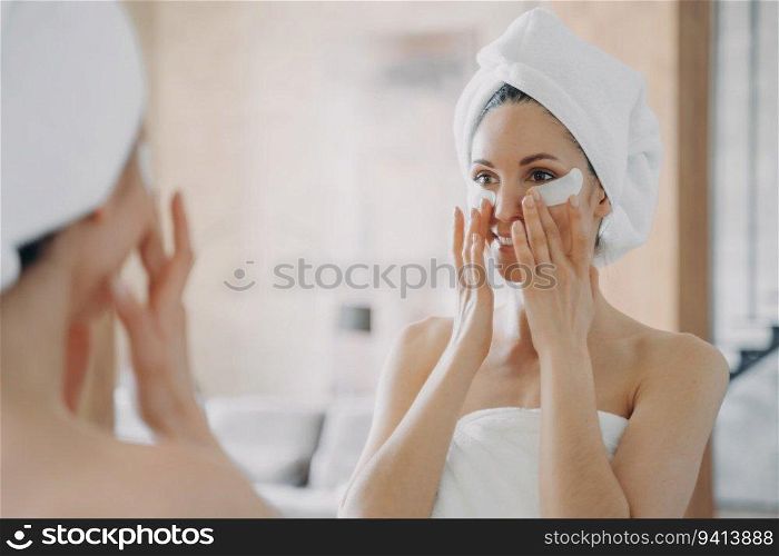 Girl applies eye patches, looks in mirror. Attractive European in towel post-bath. Young Hispanic lady showers at home. Spa relaxation, beauty routine.
