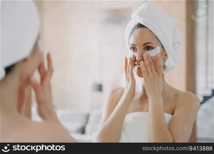 Girl applies eye patches, looks in mirror. Attractive European in towel post-bath. Young Hispanic lady showers at home. Spa relaxation, beauty routine.