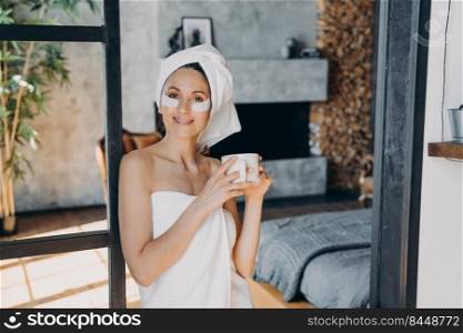 Girl applies lifting eye patches. Weekend morning beauty routine at home Young woman with cup of tea relaxing at luxurious spa resort. Attractive european woman wrapped in towel after bathing.. Girl applies lifting eye patches relaxing with cup of tea. Weekend morning at luxurious spa resort.