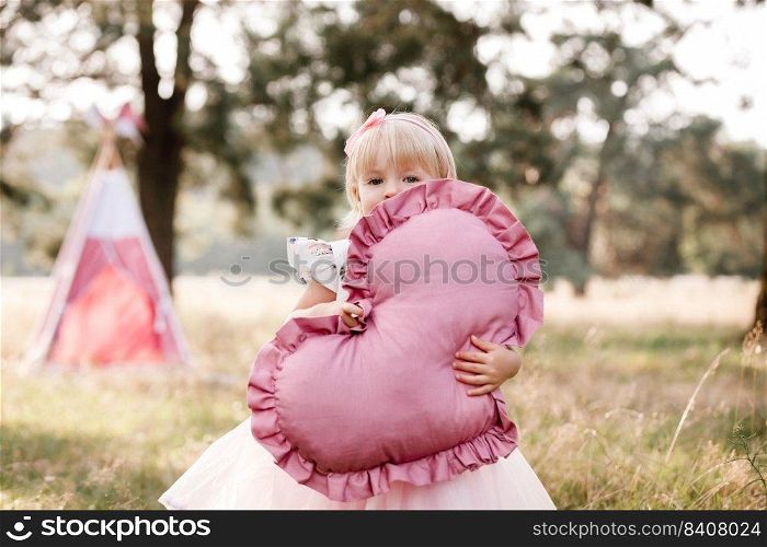Girl are dressed in pink dress and holding big pink heart in hands. Mom, dad are sitting next to wigwam decoration in the park. Family spending time outdoor in summer, having fun together. Girl are dressed in pink dress and holding big pink heart in hands. Mom, dad are sitting next to wigwam decoration in the park. Family spending time outdoor in summer, having fun together.