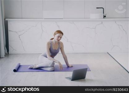 Girl at online fitness classes at home. Young smiling white woman in sportswear practicing yoga on mat by video lesson. Video call to coach. Concept of e-learning and domestic workout.. Smiling girl at online fitness classes at home. Video call to coach and workout by video lesson.