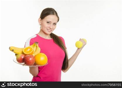 Girl athlete holding a plate of fruit and dumbbell. Young beautiful girl athlete spends Europeans physical exercise and promotes a healthy lifestyle