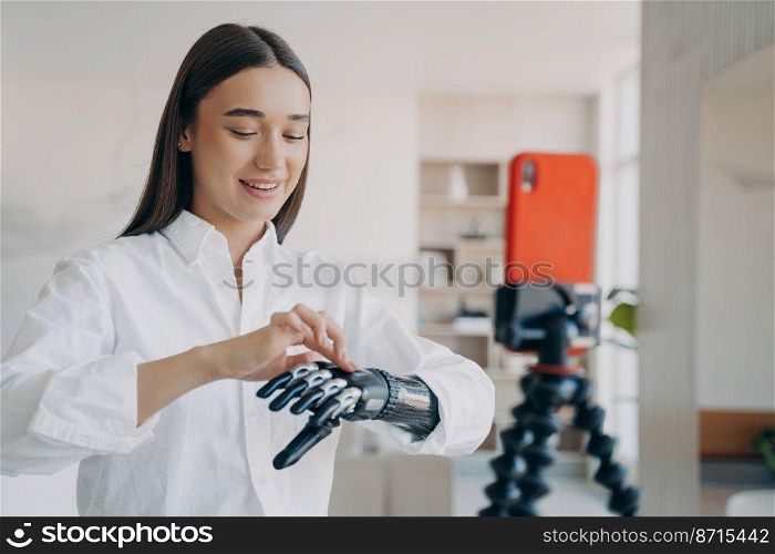 Girl blogger with disability showing bionic prosthetic arm live streaming in social networks using smartphone on tripod at home. Disabled young woman speaking to followers about artificial limb.. Blogger disabled girl shows bionic prosthetic arm live streaming in social networks using smartphone