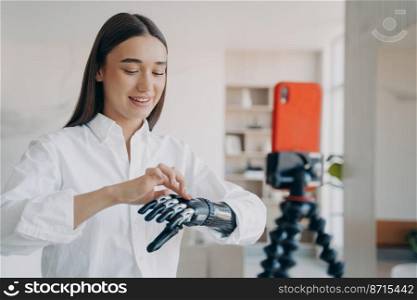 Girl blogger with disability showing bionic prosthetic arm live streaming in social networks using smartphone on tripod at home. Disabled young woman speaking to followers about artificial limb.. Blogger disabled girl shows bionic prosthetic arm live streaming in social networks using smartphone