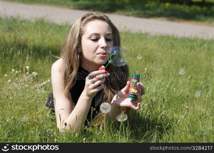 girl blowing soap bubbles lying on green grass in dandelions