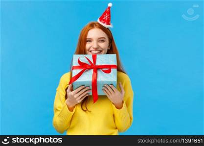 Girl cant wait give sibling her gift, smiling carry cute christmas wrapped present and wearing fancy santa hat, laughing, sharing positive emotions, attend winter holidays party, blue background.. Girl cant wait give sibling her gift, smiling carry cute christmas wrapped present and wearing fancy santa hat, laughing, sharing positive emotions, attend winter holidays party, blue background