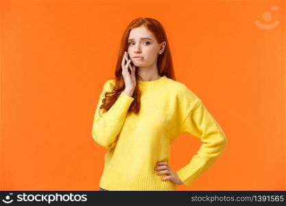 Girl catching anxiety calling someone. Perplexed or nervous cute redhead woman biting lip and looking away as having conversation on mobile phone, standing orange background sad.. Girl catching anxiety calling someone. Perplexed or nervous cute redhead woman biting lip and looking away as having conversation on mobile phone, standing orange background sad