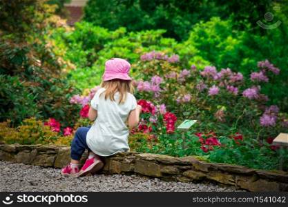 Girl child relaxing in flower garden rear view. Adorable little kid sitting around growing flowery. Young caucasian female child outside enjoy morning rest in colourful, idyllic and tranquil place.