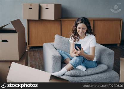 Girl clicks phone and ordering delivery service using application. Happy apartment buyer among boxes is texting on smartphone. Satisfied hispanic woman is moving to new home.. Satisfied woman is moving. Girl clicks phone and ordering delivery service using application.