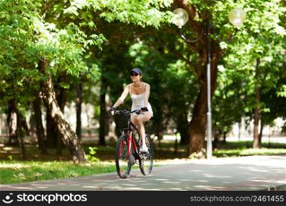 Girl cycling in the park. She&rsquo;s wearing white sport clothes, cap, her bicycle is red. It&rsquo;s a sunny summer day.