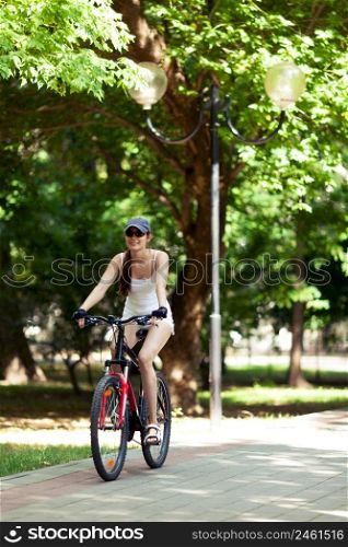 Girl cycling in the park. She&rsquo;s wearing white sport clothes, cap, her bicycle is red. It&rsquo;s a sunny summer day.
