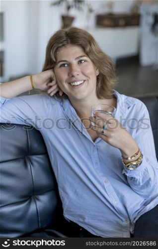Girl drinking water sitting on a couch at home and looking at camera. attractive smiling young woman sitting on couch and holding glass of water