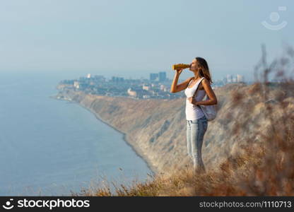 Girl drinks from a bottle of water, standing against the backdrop of a mountain and sea landscape