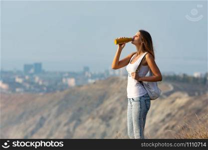 Girl drinks from a bottle of water, standing against the backdrop of a mountain and city landscape