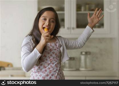Girl eating carrot standing in kitchen holding it like a singer