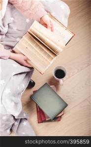 Girl enjoying the reading a book and drinking coffee at home. Young woman sitting on a chair, wrapped in blanket, holding book, relaxing at home. Portrait orientation. View from above