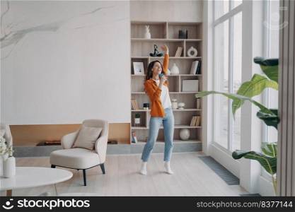 Girl having fun crazy in her living room. Happy hispanic woman is singing at home with phone as with microphone. Relaxation and dancing. Concept of emotional expressions and rebellion.. Girl having fun crazy in her living room. Relaxation and dancing at home, emotional expressions.